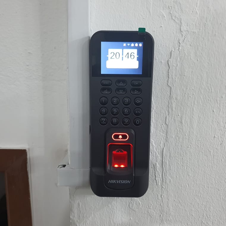 A Hikvision fingerprint terminal with a keypad and an illuminated red sensor, mounted on a white wall.