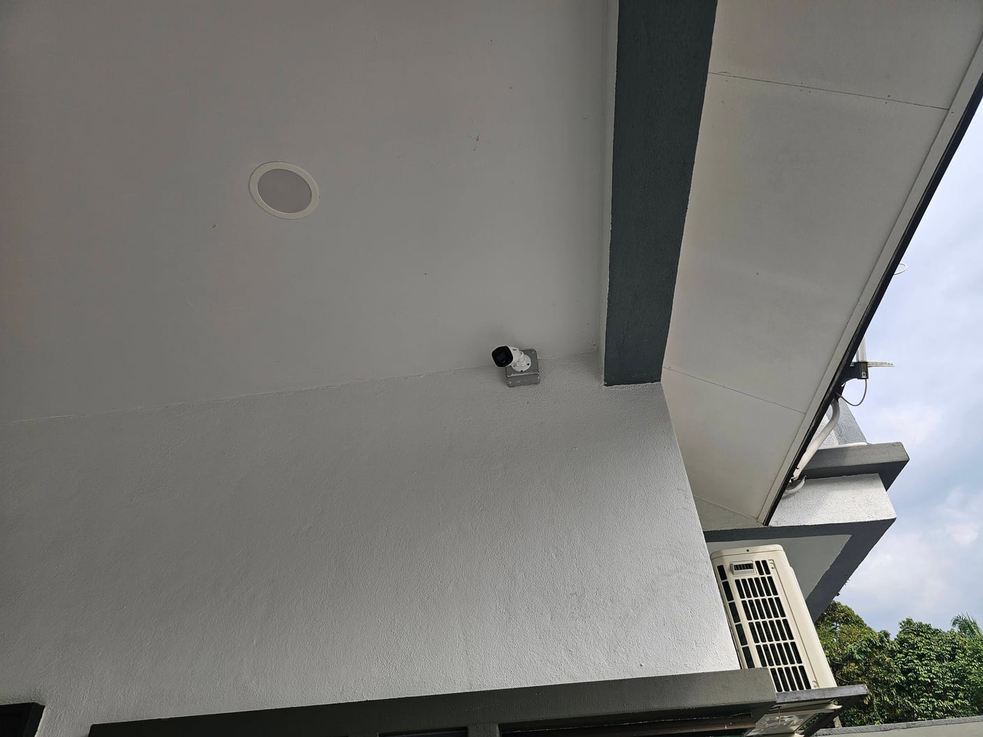 A camera on a white exterior soffit beside an air-conditioning unit.