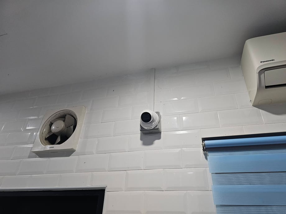 A dome camera on the ceiling of a white-tiled utility area, near a ceiling vent.